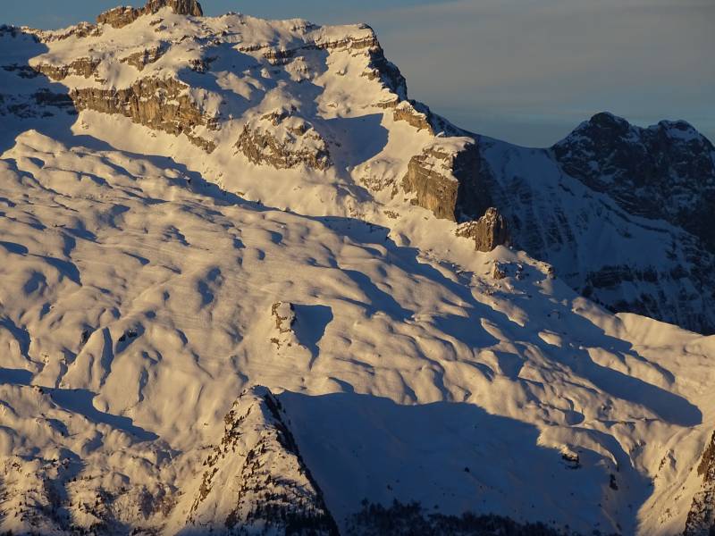 Tournette annecy alpine snowy mountain range sunset snow capped peak ski slope mountainous terrain nature evening scenery winter season The image presents a breathtaking view of a mountain landscape. Dominating the scene is a majestic snow-capped mountain, its peak reaching into the sky under a clear blue sky. Below the mountain peak, a series of smaller peaks stretch across the horizon, their surfaces blanketed in white snow. The mountain itself is covered in patches of green and brown vegetation, adding a touch of color to the otherwise monochromatic landscape. The sunlight casts an inviting glow on these areas, highlighting their textures and contrasting with the smooth, pristine snow on the peaks. In the foreground, the mountains give way to a snow-covered hill that descends gently into the distance. This hill is not as steeply sloped as the mountain behind it but still maintains an even layer of snow from top to bottom. The overall color palette of the image is dominated by shades of white and blue, with the green and brown patches on the mountains providing a pleasing contrast. The image appears to have been taken during the daytime under clear weather conditions, as evidenced by the bright lighting and the vivid blue sky.