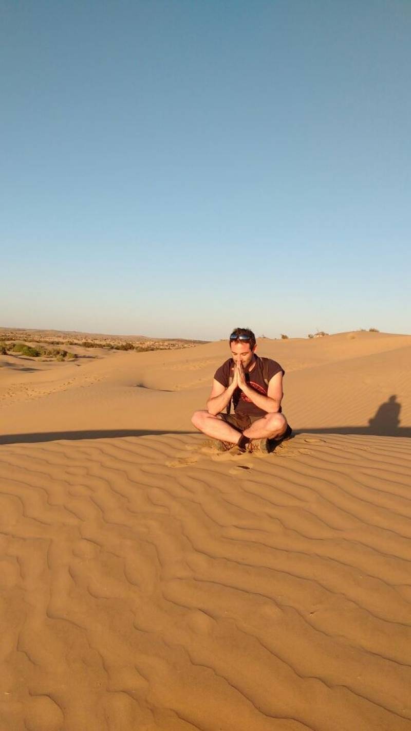 Benjamin fontaine desert posture squat voyageur dune sable priere meditation contemplatif coucher soleil solitude Une autre personne s'assied sur la sable. Elle est tournée vers la caméra et les genoux sont tirés vers leur poitrine comme si elle reposait ou réfléchissait. Elle porte également des vêtements de style informel. L'environnement entourant suggère une paysage désertique avec un ciel clair et peu de végétation.