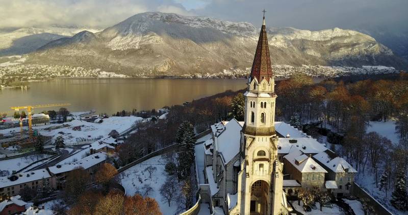 Annecy eglise montagneux rang neigeuses etaiere blanche saison neige ambiance rurale montagne enneigee architecture chretienne reflexion Le paysage montagneux pittoresque avec une église blanche dominante au centre, caractérisée par un clocher à l'arrière-plan. L'église est entourée d'un corps d'eau calme, peut-être un lac ou un ruisseau, qui reflète le paysage environnant. En arrière-plan, les sommets de montagne sont majestueux et neigés, tandis que le ciel est partiellement nuageux. Le plan-fond présente une ville avec des bâtiments et des arbres, ce qui suggère qu'il s'agit d'une partie d'une région montagnarde connue pour sa beauté naturelle. Il n'y a aucun texte visible dans l'image.