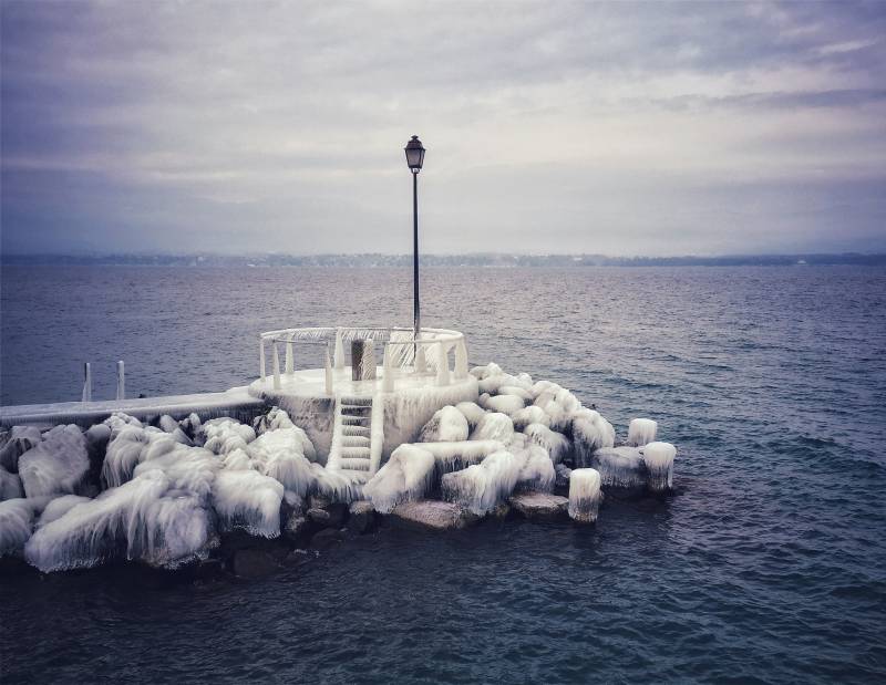 Coastline ocean pier dock foggy icy cold weather ice formation winter scenery frozen surface waterfront seaside lighthouse stormy sea coastal fog wave coastal scene. In the foreground, there is a pier with several rocks coated in what appears to be ice or frost, suggesting that the photo may have been taken during winter or early spring when freezing temperatures can cause icy accumulation on surfaces. The background features a body of water, possibly an ocean, given its vast expanse and blue-gray hues indicative of seawater. On the left side of the image, there is a small building, possibly a boathouse or storage structure for boats, with a black metal pole on top, which could serve as an anchor point or a light fixture. The sky overhead is overcast with gray clouds, and there are no visible people in the scene. The overall impression is one of a serene, cold day at a coastal location.