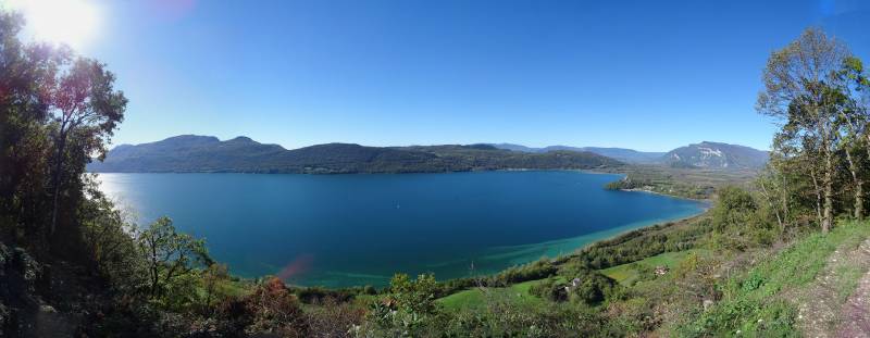 Lake scenic panoramic sunny day mountainous terrain outdoor recreation area nature tourism travel destination The image appears to show a picturesque landscape view. There is a lake or large body of water in the foreground, with clear blue waters and a calm surface. On the right side of the image, there are hills or low mountains with some vegetation visible on their slopes. Above this landscape, we can see a blue sky that suggests it might be a sunny day. In the middle ground, there is a flat area that could possibly be a small plain or an area with fewer trees. There is no text present in the image. The style of the photograph seems to capture a natural setting with scenic beauty, potentially a tourist destination or a location of interest for hiking or outdoor activities.