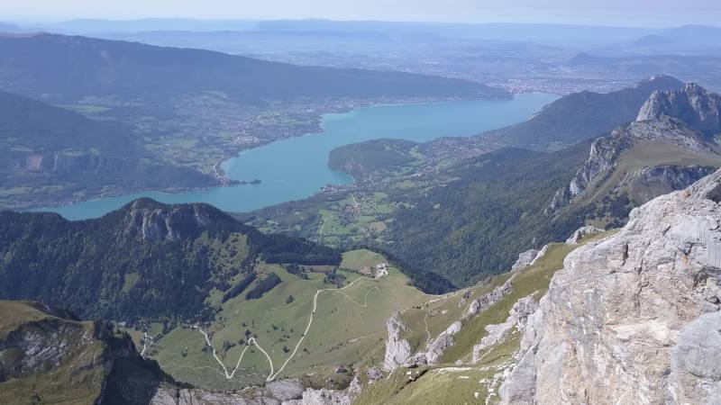 Lacs sentier randonnee panoramiques scenarios alpins tourisme naturel terre rocheuse llac montagnes destination touristique Scénique paysage présentant une région montagneuse avec un lac niché dans la vallée en dessous. Il est possible de voir clairement l'eau depuis un point d'altitude élevé, ce qui suggère que le regardeur se trouve à un altitude supérieure au lac. Les montagnes environnantes présentent des degrés de pentes variables et forment un tableau pittoresque du paysage. La verdure sur les pentes indique la présence d'arbustes ou peut-être même des forêts ou des prairies. À l'horizon, il apparaît une petite ville ou un village en bord de lac, ce qui pourrait suggérer que cette région est un lieu populaire pour les touristes ou les locaux cherchant des activités de loisirs dans la zone. Le ciel au-dessus est clair, indiquant de bonnes conditions météorologiques à l'époque du cliché. L'impression générale est celle d'une beauté naturelle et de tranquillicité.