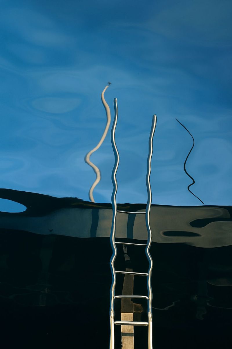 Ladder water reflection sky metal curve design pattern texture structure architecture modern minimalist geometric industrial outdoor swimming recreati metallic ladder standing vertically in the foreground, reflecting off the surface of the water. The ladder appears to be made of stainless steel, with a sleek, modern design. The water is calm and clear, reflecting the ladder and the sky above. The sky is blue with some clouds, and the reflection of the ladder and sky in the water creates a visually striking contrast. The ladder's reflection in the water appears distorted, with the lines of the ladder bending and curving in a way that suggests movement. The overall effect is one of tranquility and beauty, with the metallic ladder standing out against the natural  of the water and sky.