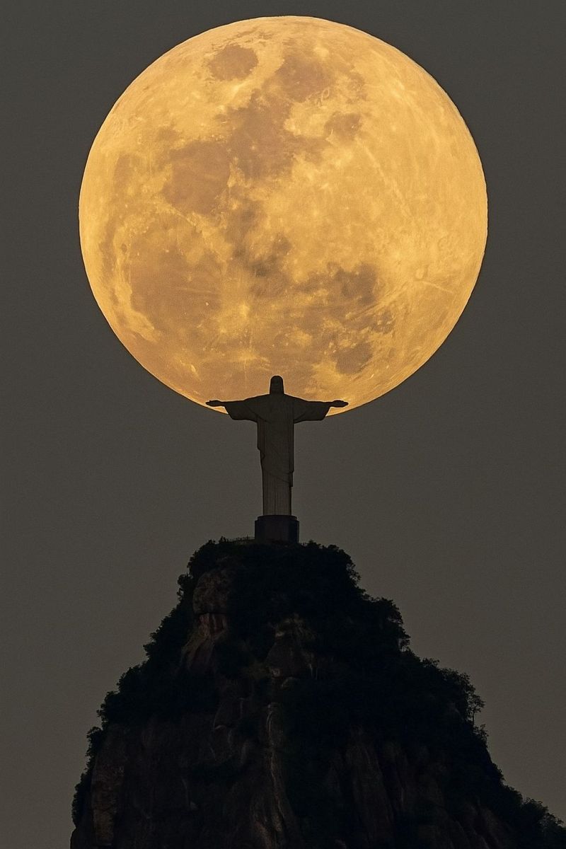 Moon supermoon christ redeemer statue rio brazil night large, full moon positioned directly above the iconic Christ the Redeemer statue in Rio de Janeiro, Brazil. The moon is bright and full, casting a golden glow over the scene. The statue, with its arms outstretched, stands on top of a rocky mountain peak, adding to the dramatic effect. The sky is dark, emphasizing the brightness of the moon and the statue. The overall scene is serene and majestic, capturing a moment of natural beauty and cultural significance.