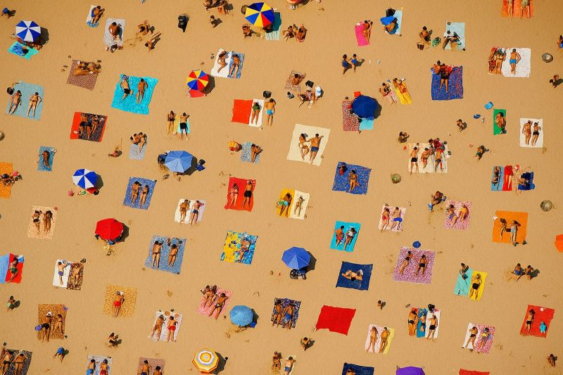 Beach people umbrella towel sunbathing crowd summer vacation relaxation outdoors sun sand recreation leisure holiday sea sky pattern aerial bustling beach scene viewed from above. Numerous people are scattered across the sandy expanse, each with their own towels and umbrellas. The beachgoers are engaged in various activities, such as sunbathing, swimming, and socializing. The towels come in a variety of colors and patterns, adding a vibrant touch to the scene. Umbrellas of different colors dot the beach, providing shade for those who prefer it. The beach is filled with a lively atmosphere, with people interacting and enjoying the sunny day.