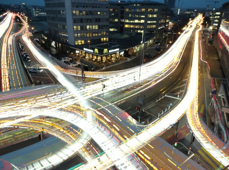 City night traffic highway cars buildings restaurant mcdonald urban intersection overpass pedestrians sidewalk construction skyscrapers cityscape fast bustling urban intersection at night, with multiple lanes of traffic flowing in various directions. The roads are illuminated by the headlights of numerous vehicles, creating a dynamic and somewhat blurry effect. The intersection is complex, with multiple flyovers and underpasses. On the left side, there is a row of parked cars, and a McDonald's restaurant is visible, with its iconic golden arches sign. The buildings in the background are tall and modern, with many windows that are lit up, indicating activity inside. The sky is dark, suggesting it is either late evening or night. The overall scene is busy and vibrant, capturing the energy of a city in motion.