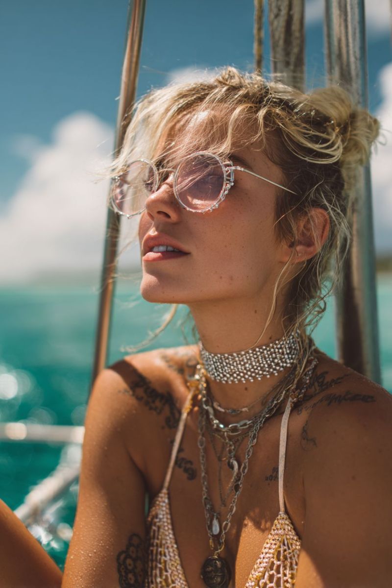 Woman sunglasses necklace boat beach ocean sun tattoo jewelry fashion accessories summer sea fashionable bikini A woman with blonde hair is standing on a boat, looking to the right. She is wearing a gold-colored bikini top with a mesh design, a silver chain necklace, and several other necklaces. She has tattoos on her arms and shoulders. She is wearing large, round sunglasses with a silver frame. The background shows a clear blue sky with some clouds and a glimpse of the ocean.
