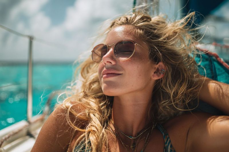 Woman boat sunglasses sun smile earring necklace ocean sky cloud sea water sunlight relaxation vacation freedom adventure lifestyle A woman with blonde hair is lying on a boat, wearing sunglasses and a necklace. She is smiling and looking to the right. The boat is sailing on the sea, and the water is calm. The sky is blue with some clouds. The woman's hair is blowing in the wind.