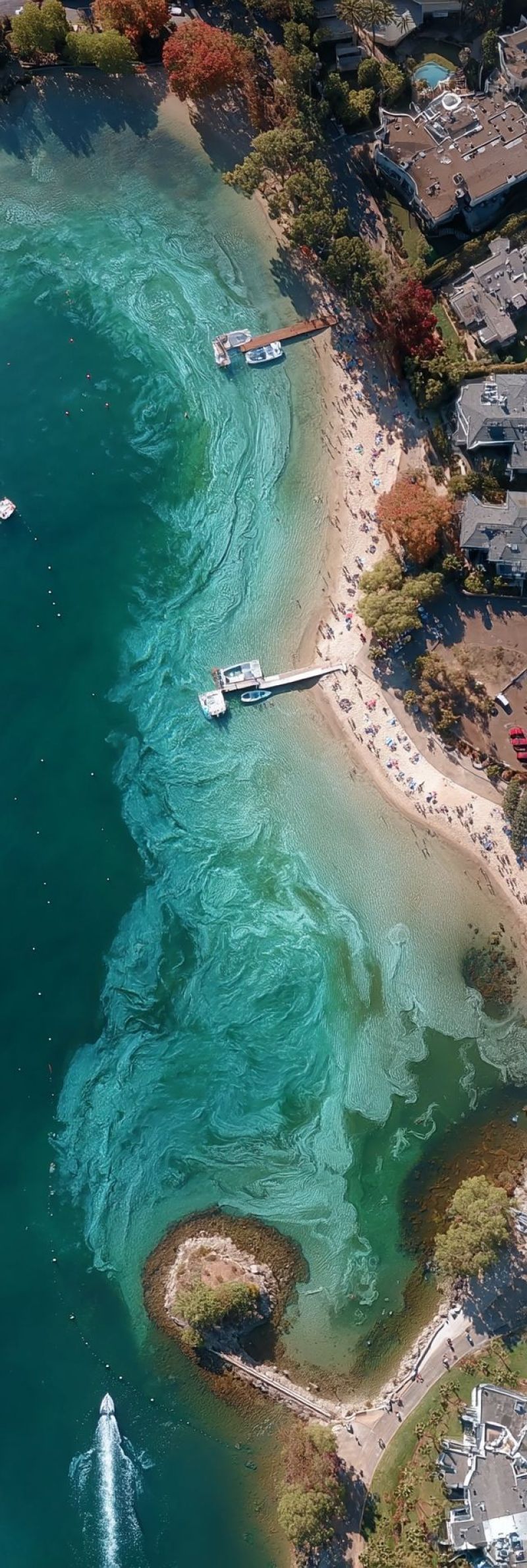 Aerien plage bleu bateau batiment ville clair cote jour quai vert maison lac ocean gens jetee reflexion riviere rivage Une vue aérienne d'une plage avec une eau bleu claire et un rivage sablonneux. L'eau a une teinte verdâtre près du rivage, possiblement à cause d'algues ou de sédiments. Il y a plusieurs bateaux amarrés le long du rivage, et un petit quai s'étend dans l'eau. La plage est peuplée de nombreuses personnes, certaines allongées sur le sable et d'autres debout. Il y a aussi plusieurs bâtiments et arbres dans la zone, dont certains ont des feuilles rouges. Le ciel est clair et bleu.