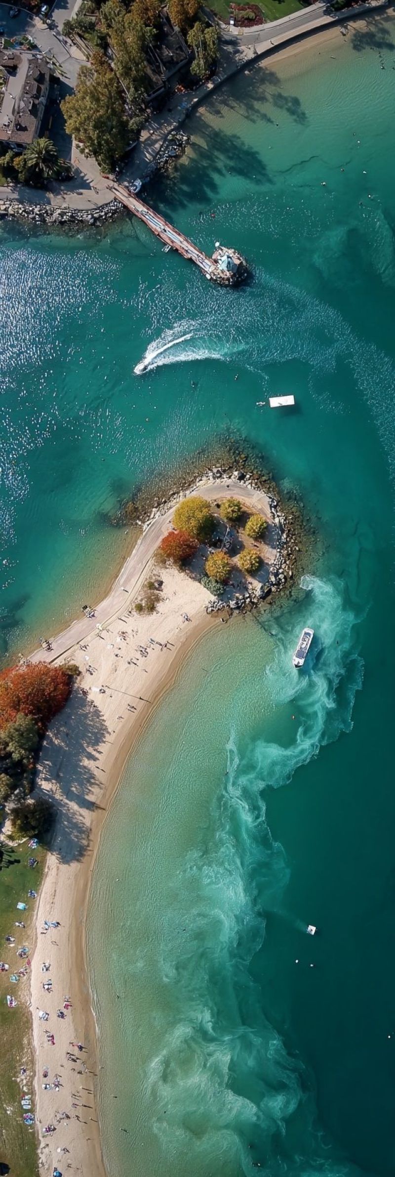 Aerial beach boat bridge building canoe city coast green island lake mountain nature outdoors people pier recreation river sea sky summer tree water An aerial view of a beach with clear blue water and a sandy shore. There are several boats in the water, including a jet ski and a small boat. The beach is surrounded by trees and bushes, and there are people sitting on the sand. In the distance, there is a bridge and a building. The water is calm, and there are no waves. The sky is clear and blue.
