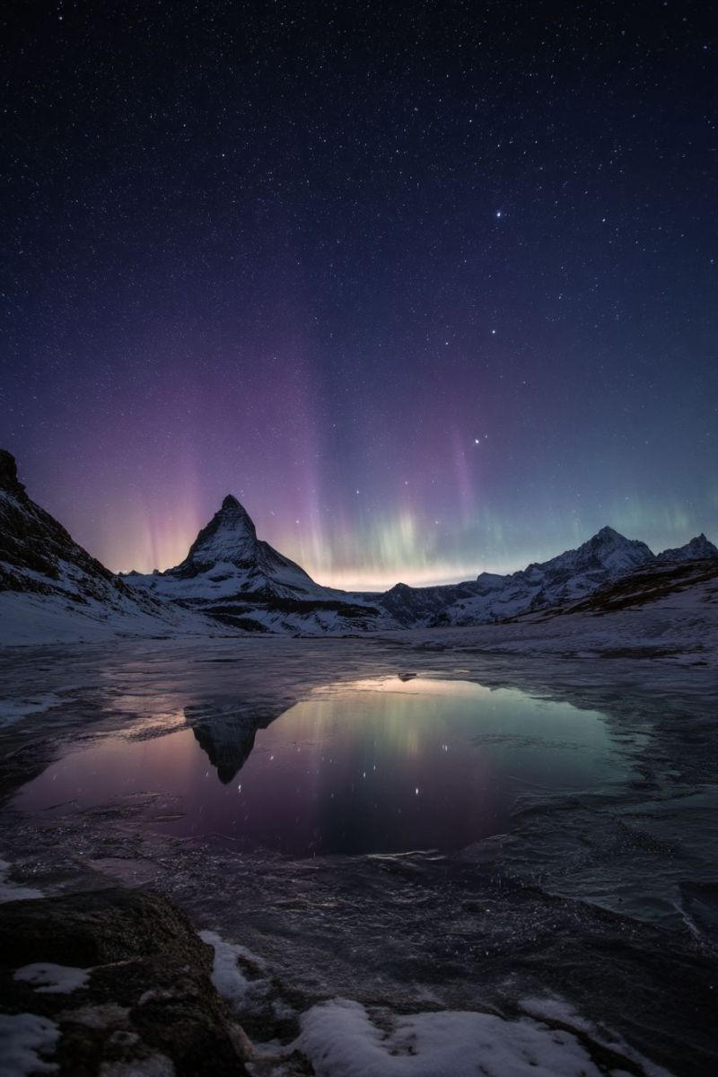 Nuit ciel montagne lac reflet neige aurore etoiles hiver glace rocher eau obscurite nuages gel froid nature Un paysage de montagne serein et majestueux est dépeint sous un ciel nocturne étoilé. Le ciel est d'un bleu profond, parsemé de nombreuses étoiles, et présente un spectacle vibrant de l'Aurore Boréale, avec des teintes de violet et de vert dansant dans le ciel. Le premier plan montre un bassin d'eau réfléchissant, miroir du ciel et des montagnes environnantes. Les montagnes sont couvertes de neige, avec des pics déchiquetés s'élevant dans le ciel. Le reflet de l'aurore et des étoiles peut être vu scintiller à la surface de l'eau, ajoutant à l'atmosphère magique de la scène.