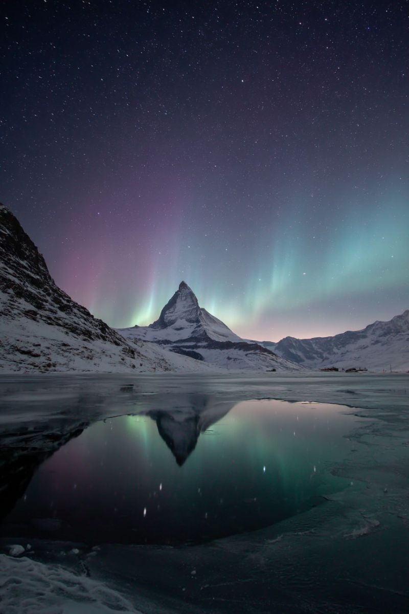Mountain sky night lake reflection aurora snow ice water star cloud dark frost winter cold frosty frozen glacier In the foreground, there is a serene, frozen lake reflecting the night sky and the majestic mountain peak. The mountain, covered in snow, stands tall and sharp against the back of the aurora borealis. The aurora lights up the sky with vibrant greens and purples, creating a magical and ethereal atmosphere. The sky is dark, dotted with stars, and the reflection of the aurora in the lake adds a touch of color to the otherwise monochromatic scene. The entire landscape is blanketed in snow, enhancing the wintery, tranquil mood of the image.