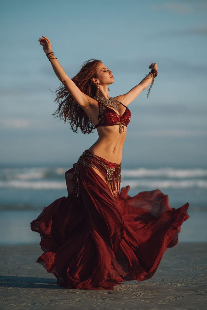 Woman beach dance costume performance jewelry earrings bracelets smiling evening sea waves sky clouds freedom belly red dress outstretched arms A woman is dancing on a beach, her arms raised high, and her body slightly bent forward. She is wearing a red belly dance costume with a matching bra and a skirt adorned with gold tassels. Her hair flows freely, and she wears gold bracelets and necklaces. The beach is sandy, and the ocean is calm in the background.