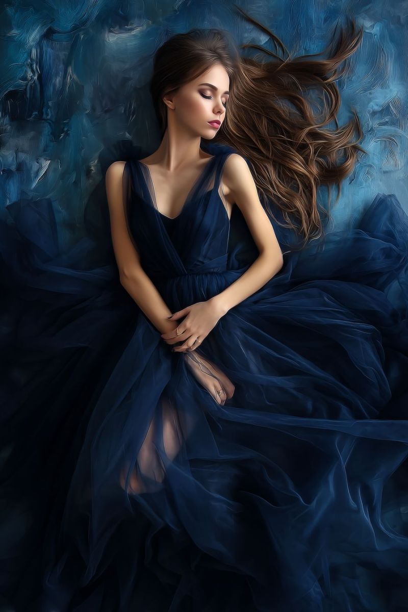 Woman fashion dress elegant beauty hair gown evening luxury glamour red carpet A woman lies gracefully on a bed, her long, wavy brown hair cascading around her. She wears a deep blue, flowing gown with a V-neckline, and her hands are gently clasped together. Her eyes are closed, and she appears to be in a serene, relaxed state. The background is a soft, abstract blue, adding to the dreamy atmosphere.