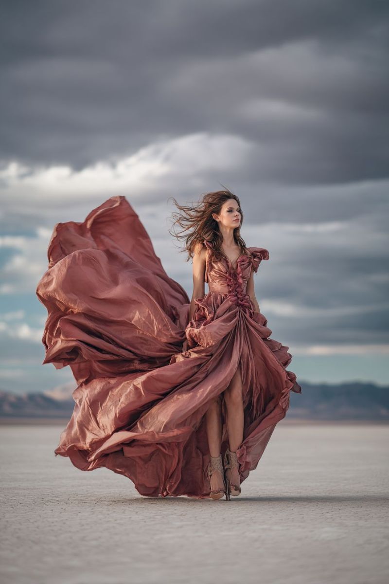 Femme robe beaute elegante soiree talon Une femme se tient sur une surface désertique plate, vaste et apparemment sans fin, sa posture gracieuse et élégante. Elle porte une robe fluide de couleur rose profond qui flotte dramatiquement autour d'elle, captant le vent et créant un sentiment de mouvement. La robe est conçue de manière complexe, avec des ruches et des plis qui ajoutent de la texture et du volume. Ses cheveux sont coiffés en vagues lâches, flottant librement dans la brise. Elle est pieds nus, avec ses pieds légèrement écartés, et son pied droit est soulevé du sol, ajoutant à la pose dynamique. L'arrière-plan présente un vaste ciel nuageux, avec la ligne d'horizon visible au loin, là où le désert rencontre le ciel. La scène générale est baignée d'une lumière douce et diffuse, possiblement d'un soleil couchant, projetant de douces ombres et mettant en valeur les contours du paysage et de la femme.