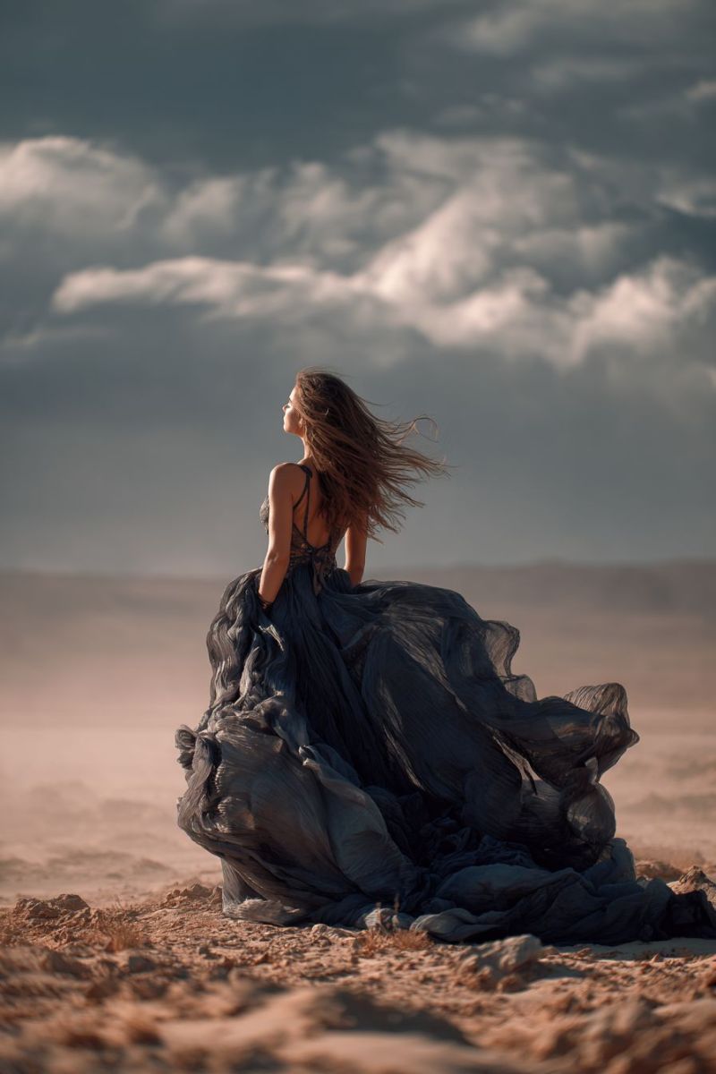 Femme robe dos vent sable ciel nuages desert beaute liberte aventure exploration nature air Une femme se tient sur une plage de sable, le dos tourné vers le spectateur. Elle porte une robe fluide bleu foncé qui flotte dans le vent, avec un décolleté halter et des détails en dentelle complexes. Ses longs cheveux ondulés sont balayés sur le côté, et elle regarde vers le haut, peut-être le ciel. L'arrière-plan présente un ciel dramatique et nuageux avec des nuances de gris et de blanc, créant une atmosphère mélancolique. Le sable est brun clair, et la scène dans son ensemble transmet un sentiment de liberté et de mouvement.