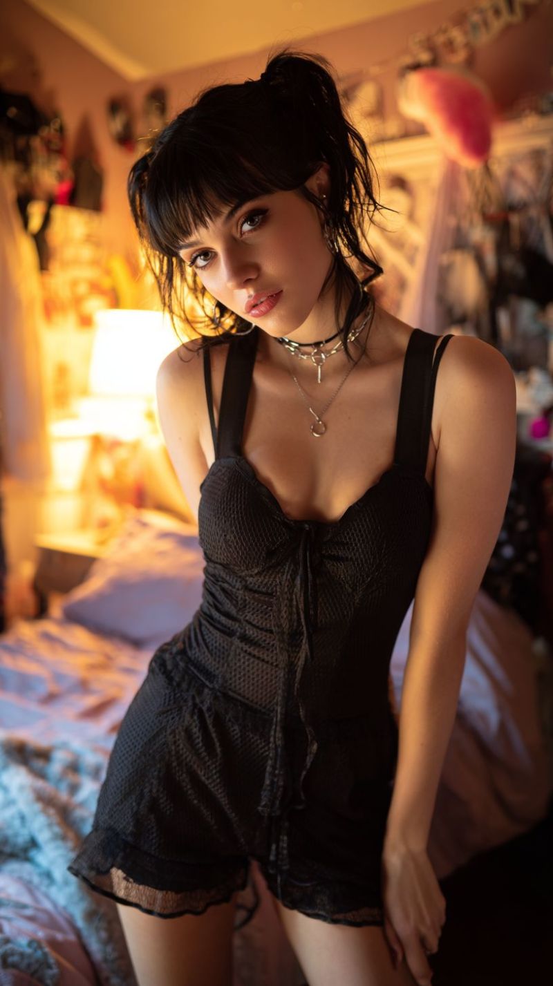 Femme piece nuit chambre coucher lingerie sommeil dormir Une femme aux cheveux courts et foncés coiffés de manière lâche et ondulée se tient dans une chambre. Elle porte un combinaison noire sans manches avec un col croisé et un petit noeud décoratif à l'avant. Sa tenue est complétée par un collier à plusieurs rangs avec un pendentif en forme de coeur. Elle pose avec une main sur la hanche et l'autre posée sur sa cuisse, regardant directement la caméra. L'arrière-plan présente un lit avec une couverture rose et une lampe sur une table de nuit, ainsi que divers objets décoratifs accrochés au mur