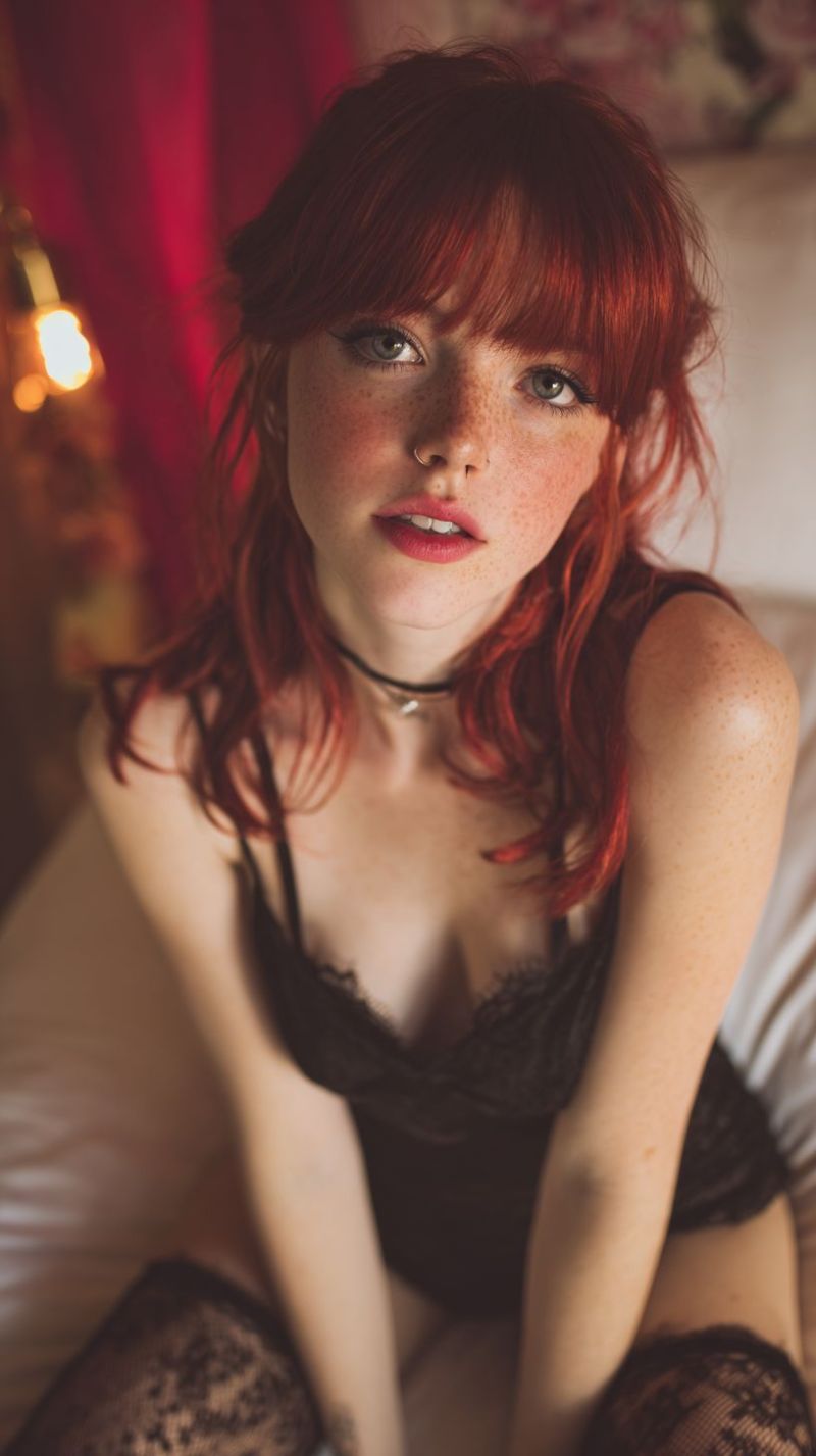 Taches rousseur piercing assis chambre coucher rouge dentelle ras cou rideau oreiller bas lingerie soutien gorge culotte jarretelle cei Une femme aux cheveux roux est assise sur un lit, portant une lingerie en dentelle noire. Elle a une expression sérieuse et regarde directement la caméra. Elle a un piercing au nez et des taches de rousseur sur le visage. L'arrière-plan est flou, avec un rideau rouge et une lumière sur le côté gauche. La lumière projette un éclairage chaleureux sur le visage de la femme.