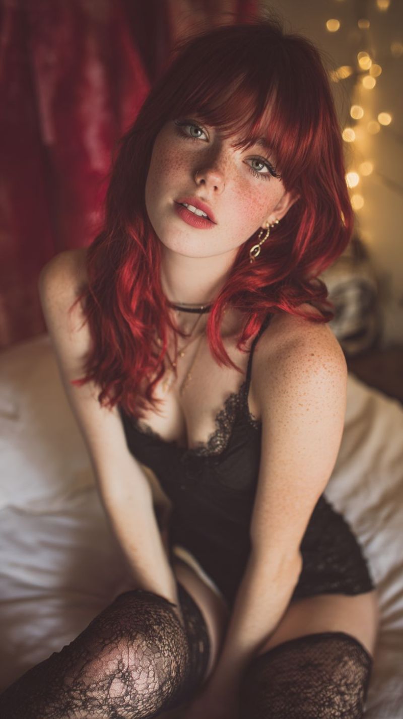 Freckles earrings necklace sitting bedroom candles smiling posing romantic intimate red lace dress blurry curtain stockings setting soft lighting A woman with red hair is sitting on a bed, wearing a black lace bra and black lace stockings. She has a serious expression and is looking directly at the camera. She has a necklace with a pendant and a ring on her finger. The background is blurry, with a red curtain and some lights.