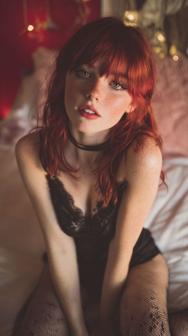 Freckles red lace choker stockings lingerie lipstick lighting wall room ambiance mood tones accents decor atmosphere A woman with red hair is sitting on a bed, wearing a black lace bra and black fishnet stockings. She has a serious expression and is looking directly at the camera. Her hair is styled in loose waves and falls over her shoulders. She is wearing a black choker necklace. The background is blurry, with a red wall and some decorative lights.