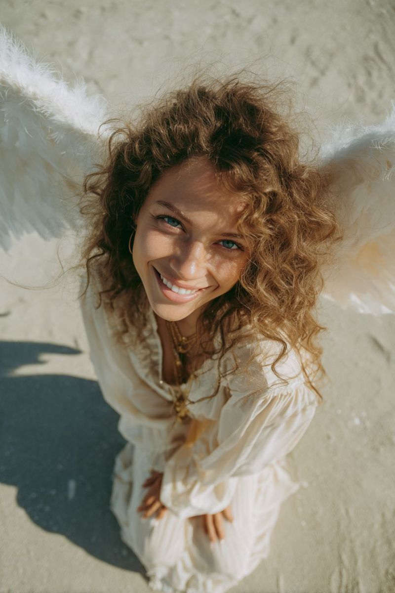 Ange ailes sourire plage sable soleil ombre oreilles collier bijoux beaute paradis divin boucle robe Une femme aux cheveux bouclés est agenouillée sur le sable, portant une robe blanche et des ailes d'ange. Elle sourit et regarde directement la caméra. Les cheveux de la femme sont soulevés par le vent, et ses ailes sont déployées derrière elle. Le sable est blanc et semble être une plage. L'ombre de la femme est projetée sur le sable.