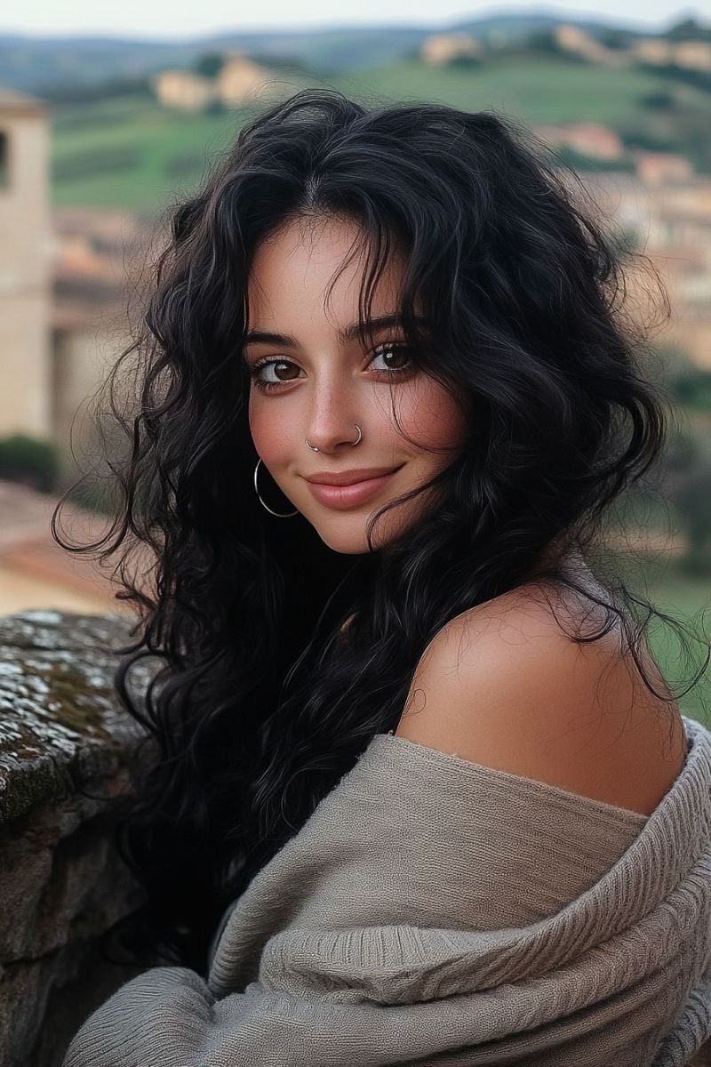 Woman outdoor travel smiling casual hair sunglasse streetwear vacation daytime fashion woman posing outdoors. She is wearing a light-colored blouse with off-the-shoulder sleeves, and her hair is styled in loose waves that cascade over her shoulders. The woman's facial features include dark eyebrows, expressive eyes, and full lips. She has a piercing on the lower lip, and her skin tone appears to be fair. Her gaze is directed towards the camera with a subtle smile. The background suggests a rural or semi-rural setting with buildings that might be houses or a small village in the distance under a clear sky. The lighting indicates either dawn or dusk, as suggested by the soft shadows on her face. There is no visible text within the image to provide additional context or information about the location, branding, or any other details.