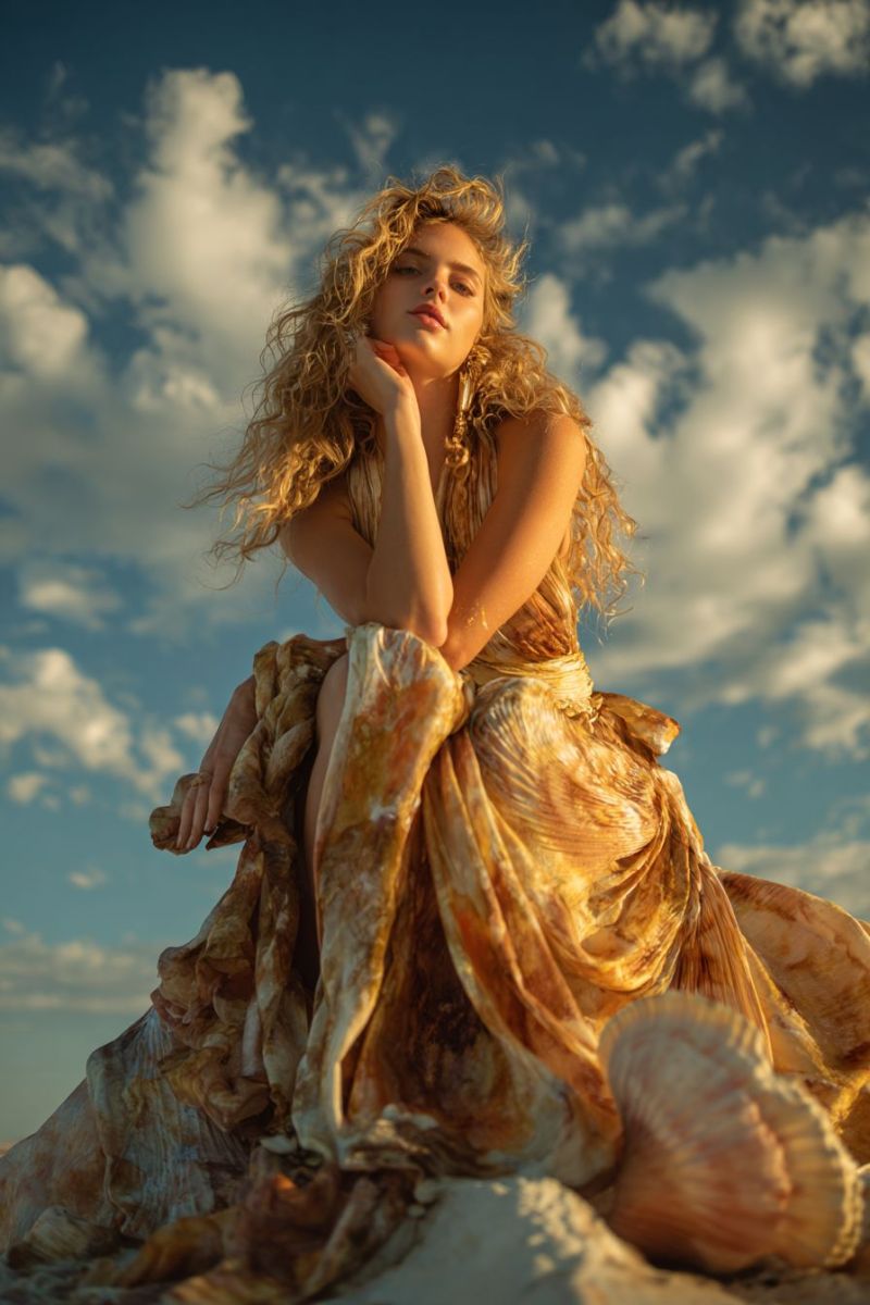 Woman dress beach seashell fashion outdoors summer elegant beauty casual curly wear chic A woman with curly blonde hair is sitting on a sandy surface, possibly a beach, with her left hand resting on her chin and her right hand on her thigh. She is wearing a flowing dress with a golden hue and a belt around her waist. The dress has a wavy pattern that resembles sea waves. The sky behind her is blue with scattered clouds. The woman's gaze is directed to the left, and she appears to be posing for a photograph.