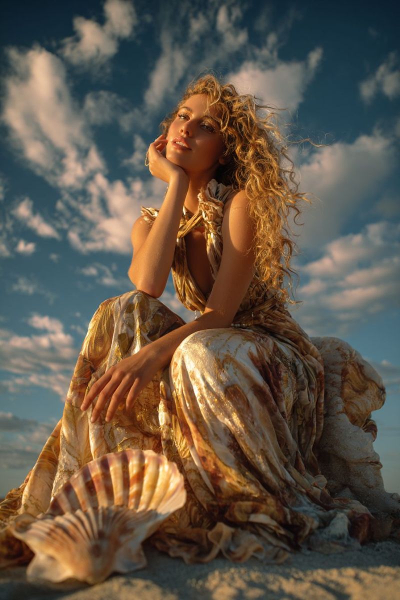 Beach woman seashell dress sitting sand sky clouds fashion outdoors summer elegance beauty jewelry accessories curly A woman with curly blonde hair is sitting on the sand, possibly on a beach. She is wearing a gold and brown dress with a leaf-like pattern. She is looking to the left, and her left hand is on her chin. She is holding a large seashell in her lap. The sky is blue with white clouds. The sunlight is shining on the woman and the seashell.