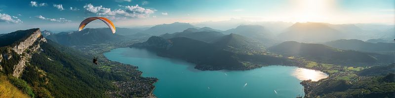 Paragliding sky mountains lake water sun clouds adventure nature outdoors travel vacation scenery horizon cliff hills trees vegetation A panoramic view of a mountainous landscape with a serene lake nestled between the peaks. The sky is a clear blue with scattered clouds, and the sun is setting, casting a warm golden light over the scene. In the foreground, a person is paragliding, their parachute a vibrant orange and white, soaring gracefully above the lake. The mountains are lush with greenery, and in the distance, a small town or village is visible, with a few buildings and a road winding through the landscape. The overall atmosphere is peaceful and picturesque, with the natural beauty of the mountains and lake creating a stunning backdrop.