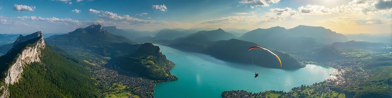 Aerien aventure falaise extreme vol planeur lac montagne parapente parapentiste ciel parachutisme Une vue aérienne d'un paysage serein présentant un grand lac tranquille entouré d'un terrain montagneux accidenté. Le lac est d'un bleu profond, reflétant le ciel bleu clair au-dessus. Les montagnes sont couvertes d'une verdure luxuriante, avec quelques falaises rocheuses visibles. Dans le ciel, une personne parapente, suspendue au-dessus du lac, avec une aile de parapente vibrante jaune et rouge. Le ciel est partiellement nuageux, avec quelques nuages dispersés à l'horizon. La scène est paisible et pittoresque, avec le parapente ajoutant un élément dynamique au cadre par ailleurs tranquille.