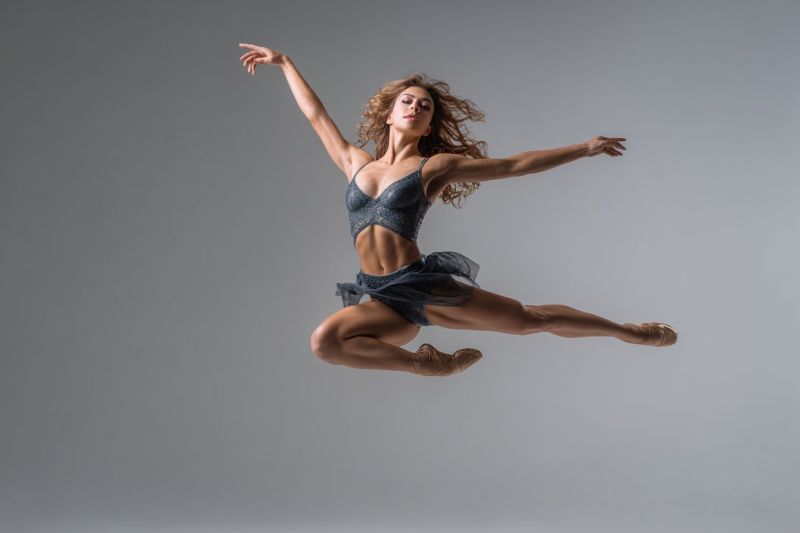 Ballet dancer dance performance grace flexibility movement expression emotion creativity stage theater choreography training technique passion inspira A woman is captured mid-air in a graceful ballet leap. She wears a fitted, dark blue leotard with a sheer, matching skirt that flows around her waist. Her arms are extended outwards, and her legs are bent at the knees, with one foot pointed and the other extended behind her. Her hair is loose and flowing, and she has a focused expression on her face. The background is a plain, light gray, providing a simple and elegant contrast to her dynamic pose.