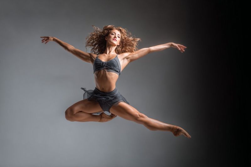 Dance ballet performance dancer jump grace flexibility expression motion movement elegant athletic fitness training studio choreography passion A woman is mid-air in a dynamic dance pose, her body forming a graceful arc. She wears a black leotard and a matching tutu, her hair flowing freely. Her arms are extended outwards, and her legs are bent at the knees, with one foot pointing upwards and the other extended behind her. Her facial expression is focused, and she appears to be performing a ballet move. The background is a solid, dark gray, providing a stark contrast to her figure.