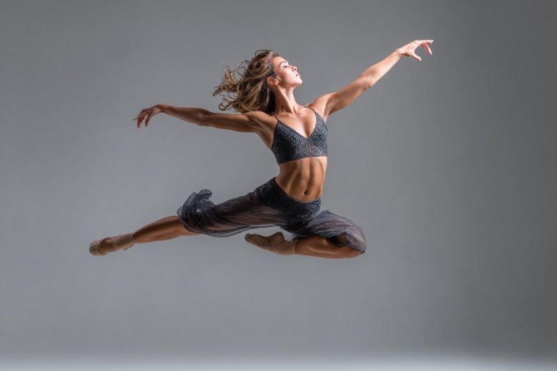 Ballet danseur performance grace expression emotion beaute flexibilite force technique entrainement passion devouement creativite imagination inspirat Une femme est capturée en plein vol dans une pose de danse dynamique. Elle porte un maillot de corps noir avec un design en dentelle et une jupe assortie. Ses bras sont étendus vers l'extérieur, et ses jambes sont pliées aux genoux, avec un pied pointant vers le haut et l'autre étendu derrière elle. Ses cheveux flottent librement, et elle regarde vers le haut, peut-être vers le plafond. Le fond est uni, d'un gris clair, offrant un contraste saisissant avec sa tenue de danse vibrante.