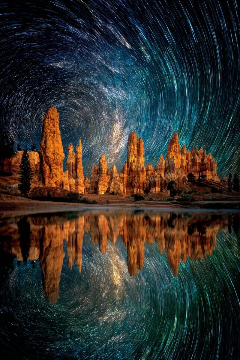 Reflexion eau nature air montagnes arbres obscurite astrophotographie capture beaute aventure nuit ciel etoile sentiers roche formations temps time Scène nocturne époustouflante mettant en vedette une série de hautes formations rocheuses orange sur fond de ciel sombre rempli d'étoiles. Les roches sont illuminées par la douce lueur des étoiles, créant un contraste saisissant. Le ciel est rempli de traînées d'étoiles, indiquant une prise de vue à longue exposition. Les reflets des roches et des étoiles sont parfaitement miroités sur l'eau calme en contrebas, renforçant l'atmosphère sereine et mystique. Le premier plan est sombre, avec seulement les contours faibles des arbres et des buissons visibles. La scène globale est une ode à la beauté naturelle et à la tranquillité, capturant l'essence d'une nuit sous les étoiles.