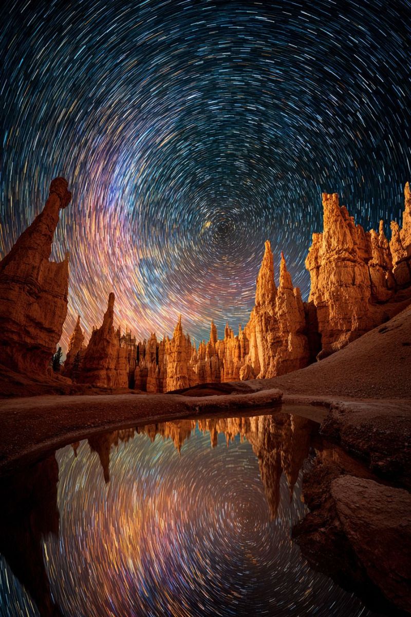 Reflexion nature air astronomie desert geologie cap montagnes rochers falaises eau ciel nuit etoile sentiers roche formations temps accelere L'image capture une scène de nuit à couper le souffle présentant un vaste paysage désertique. Au premier plan, un corps d'eau serein reflète le ciel, créant un effet de miroir. L'eau est calme, avec de douces ondulations qui reflètent les étoiles au-dessus. Le ciel est d'un bleu profond et sombre, rempli d'innombrables étoiles qui semblent tourbillonner et danser en mouvement circulaire, créant un effet fascinant de traînée d'étoiles. Les étoiles sont dispersées dans le ciel, certaines formant un arc radieux qui semble toucher l'horizon. En arrière-plan, il y a de nombreuses formations rocheuses, leurs silhouettes se détachant contre le ciel nocturne. Ces rochers sont irréguliers et variés en forme, ajoutant à la beauté sauvage du paysage. La scène globale est éclairée par la douce lueur des étoiles, projetant une lumière magique et éthérée sur l'ensemble de la scène.