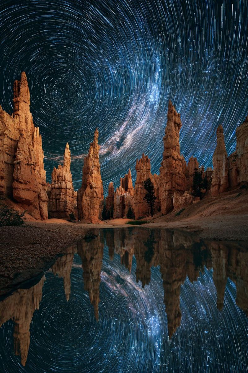 Reflection nature outdoors astronomy desert geology cape mountains trees water sky stars dark night star trails rock formations time lapse night sky filled with stars, creating a beautiful star trail effect. The foreground shows a serene landscape with a body of water reflecting the starry sky and the silhouettes of rock formations. These rock formations are tall and jagged, with a reddish-brown color, standing out against the dark sky. The water is calm, mirroring the stars and the rock formations. The sky is a deep blue, and the stars are scattered throughout, with some appearing as streaks due to the long exposure. The overall scene is peaceful and majestic, capturing the beauty of nature under the night sky.
