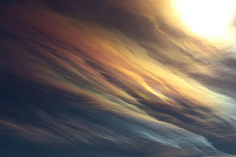 Sky clouds atmosphere rainbow weather nature colorful reflection sunlight horizon outdoors daytime brightness hue shade pattern texture The image showcases a dramatic sky filled with thick, billowing clouds. The clouds are primarily dark and gray, with hints of orange and yellow, suggesting the presence of sunlight breaking through. A vivid, multicolored arc, likely a rainbow, is visible, stretching across the sky. The sun, though not directly visible, is implied by the light illuminating the clouds and creating the rainbow effect. The overall scene is dynamic, with the clouds appearing to move and swirl, creating a sense of depth and motion.