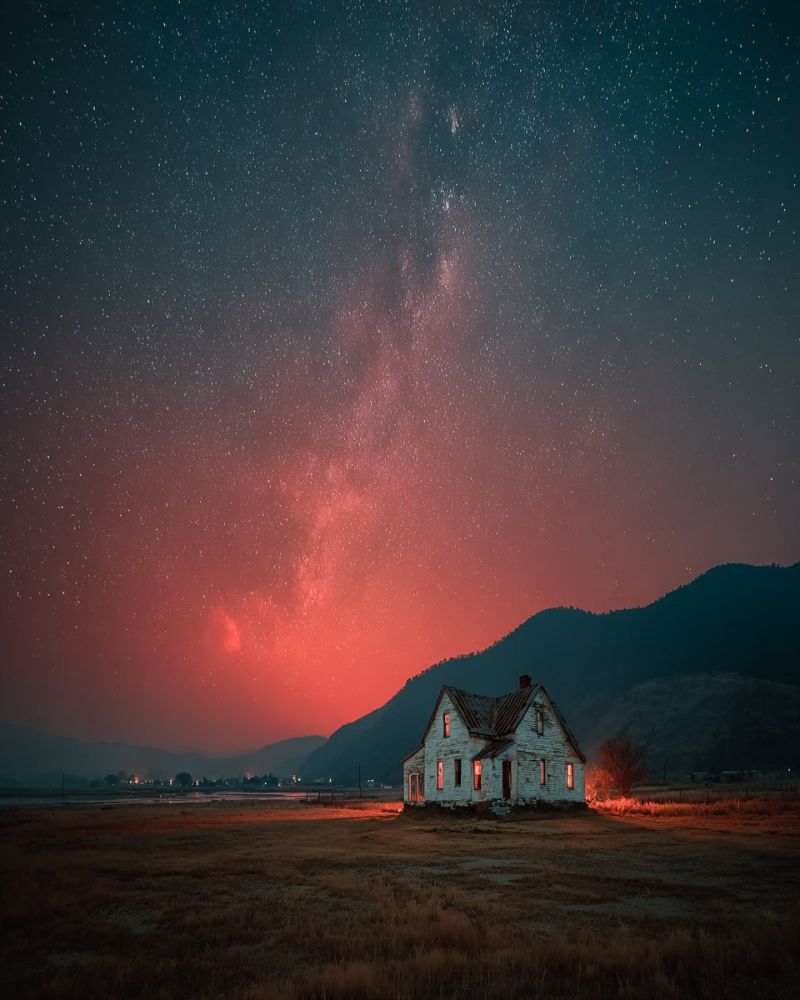 Maison nuit ciel etoiles montagne champ architecture batiment toit fenetre porte rouge reflet ombre herbe arbres voie lactee Au centre de l'image, il y a une vieille maison usée se tenant seule dans un vaste champ ouvert. La maison a un aspect usé et rustique, avec un toit en pente et une cheminée. Le ciel au-dessus est d'un bleu profond et sombre, rempli d'innombrables étoiles, et la galaxie de la Voie Lactée est visible de manière proéminente, s'étendant à travers le ciel avec une teinte rougeâtre. L'horizon est marqué par une silhouette de montagnes, ajoutant à l'atmosphère sereine et isolée. L'avant-plan est un champ d'herbe sèche, et la maison est entourée d'une lueur rougeâtre, possiblement d'un feu proche ou du soleil couchant.