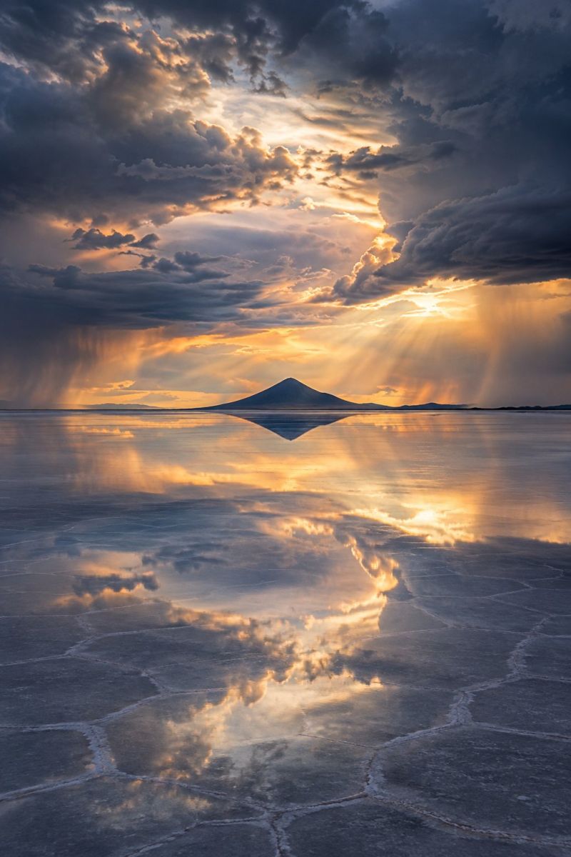 Coucher soleil reflet ciel nuages montagne lac eau horizon nature meteo atmosphere nuageux mer une vaste étendue d'eau plate, probablement une plaine de sel, sous un ciel spectaculaire. La surface de l'eau est lisse et réfléchissante, reflétant le ciel et les nuages au-dessus. Le ciel est rempli de nuages sombres et imposants, traversés par les rayons du soleil, créant un contraste saisissant. Le soleil se couche ou se lève, projetant une lumière chaude et dorée sur la scène. Au loin, la silhouette d'une montagne se détache sur l'horizon, donnant une impression d'échelle et de profondeur au paysage. L'ambiance générale est à la fois sereine et dramatique, les jeux d'ombres et de lumières accentuant la beauté de la scène.