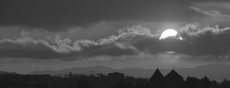 Annecy chateau soleil nuages ciel montagnes batiments architecture urbain construction grue arbres foret collines horizon meteo atmosphere ombres Une photographie en noir et blanc montre un ciel dramatique rempli de nuages épais et sombres. Le soleil est partiellement obscurci par les nuages, créant un effet de silhouette. La lumière du soleil traverse les nuages, projetant une douce lueur sur le paysage en contrebas. Au premier plan, il y a des silhouettes de bâtiments, d'arbres et d'une grue, suggérant un cadre urbain ou industriel. Les montagnes lointaines sont à peine visibles, ajoutant au sentiment de profondeur et d'échelle. L'ambiance générale est sombre et atmosphérique, avec l'interaction entre la lumière et l'ombre créant un contraste visuel saisissant.