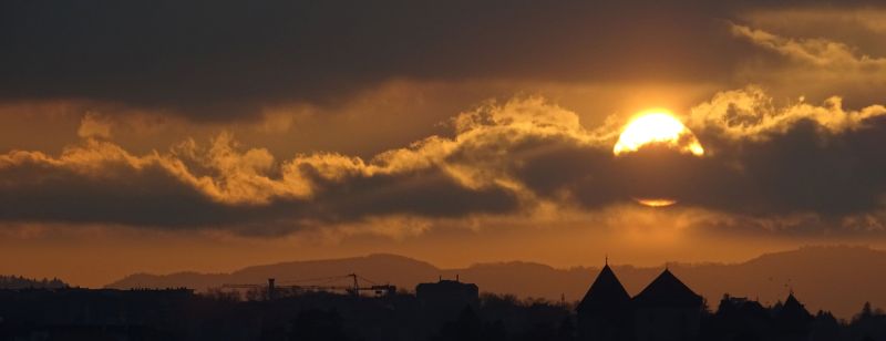 Annecy castle sunset sky clouds sun mountains cityscape architecture buildings town hills trees orange yellow pink dark night evening sunset over a landscape. The sun is partially obscured by clouds, creating a dramatic effect as it shines through gaps in the cloud cover. The sky is a mix of deep oranges and purples, indicating the time of day is late afternoon or early evening. Below the sky, there are silhouettes of buildings and trees, suggesting a town or city. In the distance, there are hills or mountains, adding depth to the scene. The overall mood is serene and picturesque, with the warm colors of the sunset casting a golden glow over the landscape.