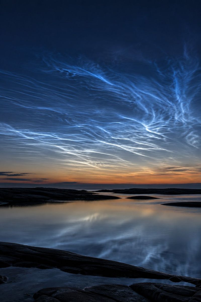 Sky clouds water reflection sunset nature horizon rocks sea shore evening blue orange dark calm quiet peaceful beauty serene twilight scene with a deep blue sky transitioning into a warm orange and pink horizon. The sky is adorned with wispy, illuminated clouds that appear to be noctilucent clouds, glowing faintly in the fading light. These clouds are scattered across the sky, creating a delicate and ethereal pattern. Below, a calm body of water reflects the sky's colors, with gentle ripples adding to the tranquil atmosphere. In the foreground, there are dark, rocky outcrops that jut out into the water, their edges softly illuminated by the setting sun. The overall scene is peaceful and picturesque, capturing the beauty of a natural landscape at dusk.
