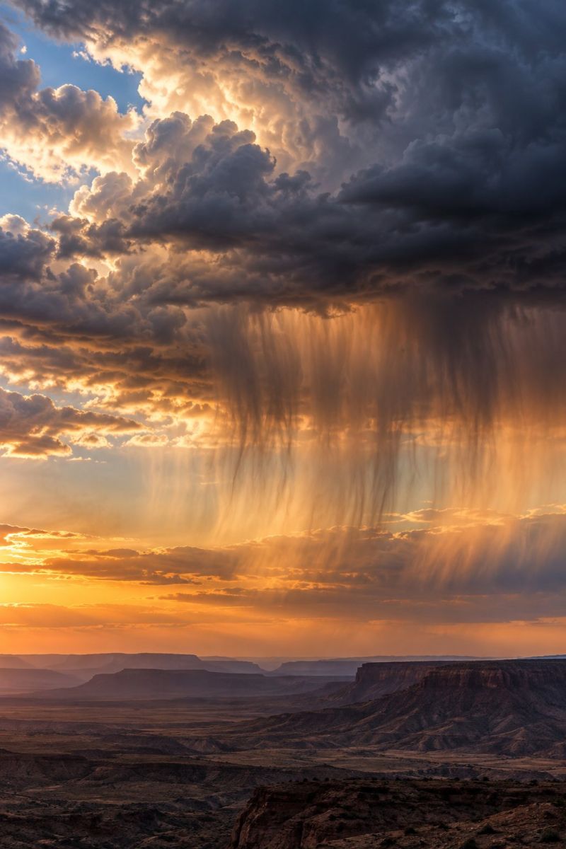Sky clouds sun sunset mountains desert weather nature outdoors horizon atmosphere rain storm valley rocks geology The image captures a dramatic sunset over a vast desert landscape. The sky is filled with towering, dark clouds, illuminated from below by the setting sun. The sun's rays pierce through the clouds, casting a golden glow on the landscape below. The clouds appear to be raining, with raindrops visible as they fall. The desert below is vast and rugged, with rocky outcrops and a horizon that stretches far into the distance. The overall scene is one of dramatic beauty, with the interplay of light and shadow creating a striking visual effect.
