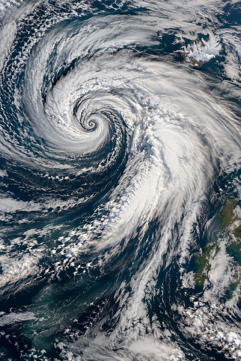 Atmosphere nuages cyclone terre environnement brouillard geographie ouragan glace meteorologie ocean satellite mer tempete climat extreme geographique vue satellite d'un puissant système de tempêtes tourbillonnant au-dessus de l'océan. La tempête a une forme en spirale distincte avec des nuages blancs formant un motif dense et tourbillonnant. Le centre de la tempête apparaît plus sombre, ce qui indique une zone de basse pression. L'océan environnant est d'un bleu profond, avec des nuages bleus et blancs plus clairs éparpillés autour de la tempête. En arrière-plan, des traces de masses continentales, peut-être des îles ou des côtes, sont visibles à travers les nuages. La scène d'ensemble est dynamique et dramatique, mettant en valeur l'immense puissance et la beauté d'un phénomène météorologique naturel.