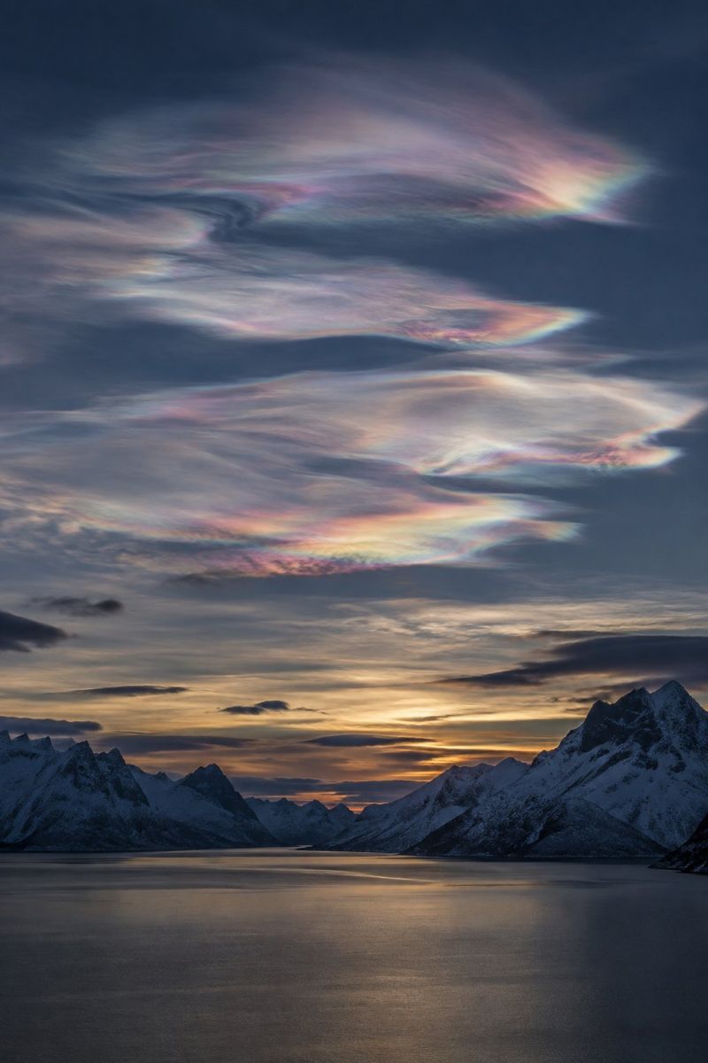 Ciel nuages montagnes lac coucher soleil reflet eau neige nature horizon atmosphere beaute air paysage serein et spectaculaire au coucher du soleil. Le ciel est rempli de nuages hauts et vaporeux qui présentent un spectre de couleurs, notamment des roses, des violets et des bleus, suggérant la présence de nuages nocturnes. Ces nuages sont positionnés au-dessus d'une vaste étendue d'eau calme, peut-être un fjord ou un lac, reflétant les couleurs du ciel. Au premier plan, des montagnes enneigées avec des sommets acérés ajoutent à l'atmosphère majestueuse et tranquille. L'horizon est peint avec les teintes chaudes et dorées du soleil couchant, projetant une douce lueur sur l'ensemble de la scène.