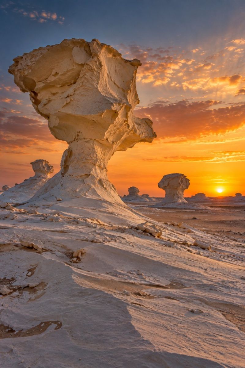 Desert sunset sand sky clouds sun nature outdoors geology rock sunrise horizon shadow beauty scenery formation In a vast desert landscape, a large, uniquely shaped rock formation stands prominently against the back of a setting sun. The rock, with its irregular, almost organic contours, casts a long shadow onto the sandy terrain below. The sky is awash with warm hues of orange and pink, transitioning into deeper blues as the sun dips lower. Smaller, similarly shaped rock formations are scattered around, adding to the surreal and otherworldly atmosphere. The scene is bathed in the soft, golden light of the sunset, creating a serene and almost magical ambiance.