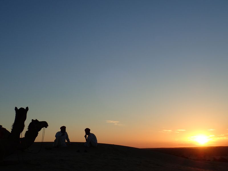 Desert sunset camel sky horizon people sitting mountain travel adventure nature outdoors backlit reflection warm evening distant two people are sitting on the ground in a desert landscape, silhouetted against the setting sun. The sun is low on the horizon, casting a warm, golden light across the scene. The sky transitions from a deep blue at the top to a lighter blue and then to a soft orange and pink near the horizon. The ground is sandy, with small dunes visible in the distance. The two individuals are facing each other, engaged in conversation. One person is wearing a hat, and both are dressed in long, flowing garments. Nearby, a camel is partially visible, its head turned towards the setting sun. The overall mood is serene and tranquil, with the vast, open desert stretching out into the distance.