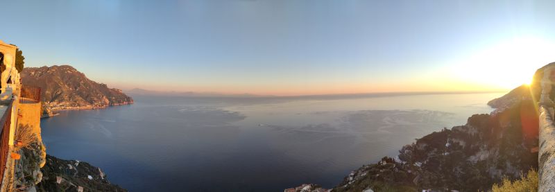 Infinito ravello cimbrone amalfi sunset panoramic From a high vantage point, the image captures a breathtaking sunset over a vast expanse of the Mediterranean Sea. The horizon is a gradient of warm colors, transitioning from a deep orange near the sun to a soft pink and purple further away. The sea is calm, with gentle ripples reflecting the colors of the sky. On the left side, a rocky cliff with a small structure, possibly a watchtower or a small building, is perched on the edge. The cliff is surrounded by lush greenery, and in the distance, another landmass is visible, possibly an island. The sky is clear, with a few scattered clouds, and the sun is partially obscured by the horizon, casting a golden glow over the entire scene.