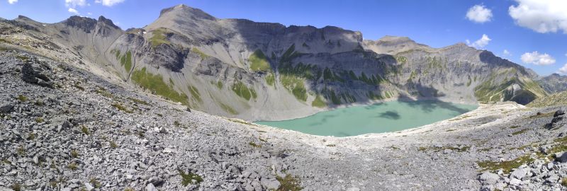 Lac emosson cheval panoramic panoramic view of a mountainous landscape. In the foreground, there is a rocky terrain with scattered vegetation. The rocks are jagged and uneven, creating a rugged appearance. Moving towards the center, there is a serene lake nestled within the mountains. The lake is surrounded by a mix of rocky cliffs and grassy areas, with patches of green vegetation adding a touch of color to the scene. The mountains rise majestically in the background, their peaks covered in snow, creating a striking contrast against the clear blue sky. The sky is dotted with a few fluffy clouds, adding to the picturesque beauty of the landscape.