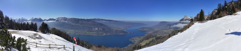 Parapent deco forclaz annecy D'un point de vue élevé, une vue panoramique d'un paysage montagneux enneigé se dévoile. L'avant-plan est recouvert de neige, avec une clôture en bois marquant le bord d'un chemin. À gauche, un petit groupe d'arbres persistants se dresse contre le fond enneigé. Le centre de l'image est un vaste lac serein, dont la surface reflète le ciel bleu clair au-dessus. Au-delà du lac, une série de collines et de montagnes ondulantes s'étendent jusqu'à l'horizon, leurs sommets saupoudrés de neige. Le ciel est d'un bleu brillant, avec quelques filaments de nuages dispersés à travers lui. Toute la scène est baignée dans la douce lumière d'une journée ensoleillée, créant un paysage hivernal paisible et pittoresque.